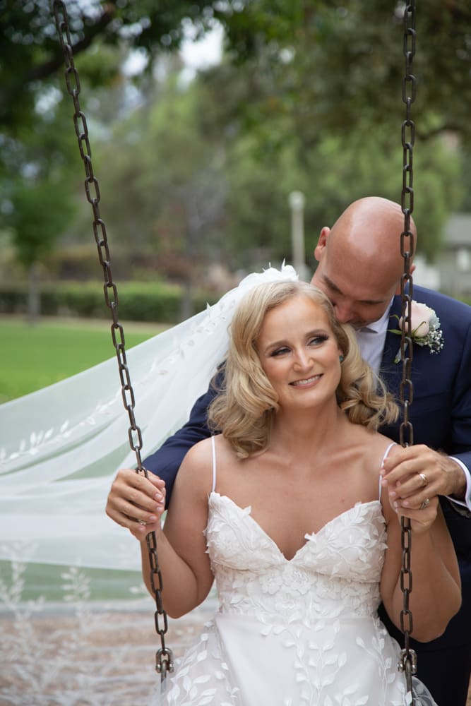 A bride in a white dress and veil sits on a swing smiling, while the groom in a dark suit stands behind her, gently holding her and nuzzling her head.